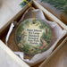 A round ornament with a bird and tropical plants, placed in a box with white tissue paper.