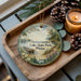 A decorative ornament featuring a serene lake scene with a boat, pine trees, and a cabin, placed on a wooden tray alongside pine cones and a lit candle.