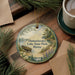 A decorative ornament featuring a lake scene with a boat, surrounded by pine trees and flowers, placed on a wooden surface next to a cup of coffee.