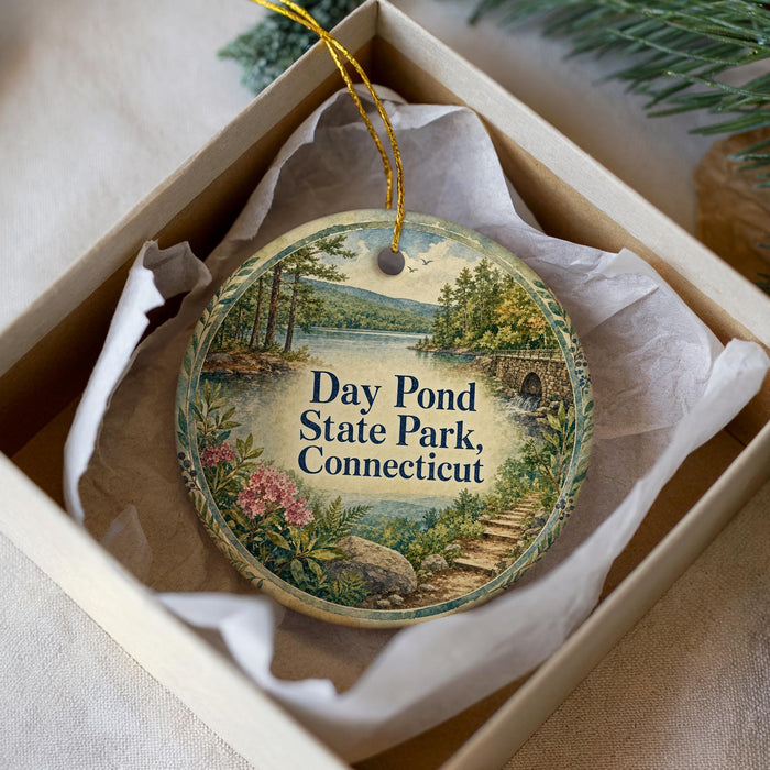 A decorative ornament in a box, featuring a scenic landscape with a pond, trees, and a bridge.