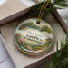 A decorative ornament in a box, featuring a scenic landscape with a pond and trees, with the text "Day Pond State Park Connecticut" printed on it.