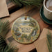 A decorative ornament featuring a scenic image of Deep Creek Lake State Park, Maryland, with a boat and a waterfall in the background. The ornament is hanging on a wooden surface, surrounded by pine branches and a cup of coffee.