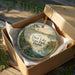 A decorative ornament in a brown box, featuring a scenic landscape with a lake, palm trees, and wildlife.