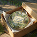 A decorative ornament in a box, featuring a scenic landscape with a castle, trees, and a lake.