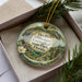 A decorative ornament in a box, featuring a scenic landscape with palm trees, mountains, and a bird.