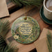 A decorative ornament with a floral design hangs on a wooden surface, surrounded by pine branches and a cup of coffee.