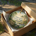 A round ornament in a box, featuring a scenic landscape with a river, trees, and a bridge, with the text "Dinosaurs State Park, Connecticut" printed on it.