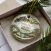 A decorative ornament in a box, featuring a scenic landscape with a river, trees, and a house.