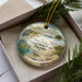 A decorative ornament in a box, featuring a beach scene with a bird, turtle, and palm tree.