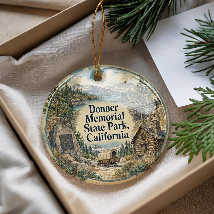 A decorative ornament with a scenic landscape of Donner Memorial State Park, California, featuring a wagon and a cabin, is displayed in a box with pine branches.