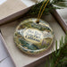 A decorative ornament with a landscape scene featuring a river, mountains, and a house, placed in a box with pine branches.