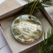 A decorative ornament with a beach scene and a lighthouse, placed in a box with pine branches.
