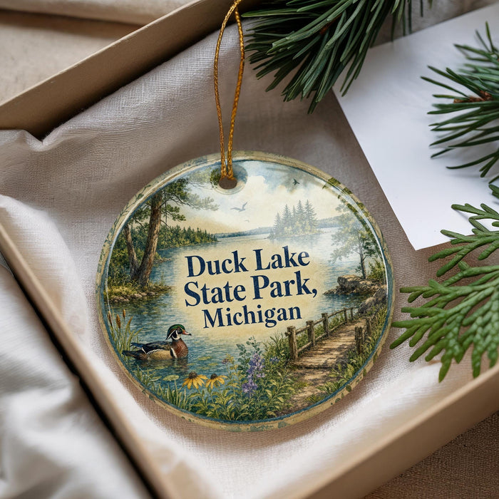 A decorative ornament with a scenic landscape featuring a lake, trees, and a path, hanging from a string.