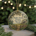 A round ornament with a painting of a farm and a windmill, surrounded by greenery, placed on a surface with a blurred background of lights.