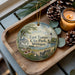 A decorative ornament with a scenic view of East Tawas City Park, Michigan, featuring a gazebo, bench, and flowers, along with pine cones and a lit candle.