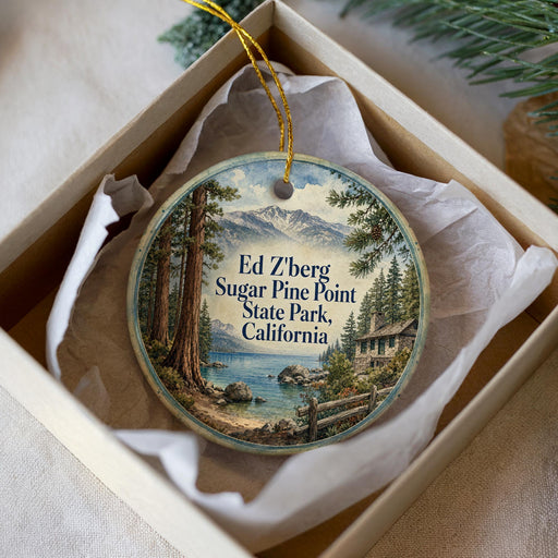 A decorative ornament in a white box, featuring a scenic landscape with a lake, mountains, and pine trees.