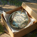A round ornament in a box, featuring a scenic image of Ed Zberg Sugar Pine Point State Park, California.