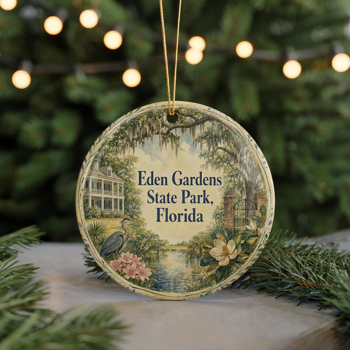 A round ornament with a painting of a house and trees, surrounded by greenery, hangs in front of a blurred Christmas tree with lights.
