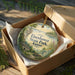 A decorative ornament in a box, featuring a scenic landscape with a house, trees, and a body of water.