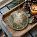 A wooden tray with a decorative ornament featuring a scenic landscape of Eldorado Canyon State Park, Colorado, along with pine cones and a lit candle.