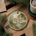 A decorative ornament with a scenic landscape of Elinor Bedell State Park, Iowa, featuring a lake, mountains, and pine trees. The ornament is hanging on a wooden surface, surrounded by pine branches and a cup of coffee.