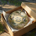 A decorative ornament in a box, featuring a scenic landscape with a deer and a waterfall, with the text "Elk City State Park, Kansas" printed on it.