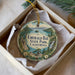 A decorative ornament in a box, featuring a scenic landscape with mountains, water, and pine trees.