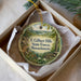 A round ornament with a scenic forest landscape, featuring a mountain, trees, and a river, is displayed in a box.