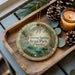 A decorative ornament with a tropical scene, featuring palm trees, a bird, and a pond, is displayed on a wooden tray alongside pine cones and a lit candle.