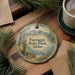 A decorative ornament with a scenic mountain landscape and pine trees, featuring the text "Farragut State Park, Idaho" on it, is displayed on a wooden surface alongside a cup of coffee and some papers.
