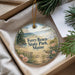 A round ornament with a scenic beach and park landscape, featuring a bird and pine trees, is displayed in a box with pine branches.