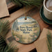A round ornament with a beach scene and text, hanging on a wooden surface next to a cup of coffee and pine branches.