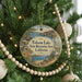 A round ornament hanging on a Christmas tree, featuring a scenic landscape with a lake, mountains, and a bird.