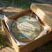 A round ornament in a box, featuring a scenic landscape with a lake, mountains, and a bird, along with the text "Folsom Lake State Recreation Area, California".