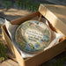 A round ornament in a box, featuring a scenic landscape with mountains, trees, and a fish, is displayed on a wooden surface.
