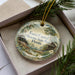 A decorative ornament in a box, featuring a scenic landscape with a house, trees, and a body of water.