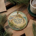 A round ornament with a painting of Fort Clinch State Park, Florida, is displayed on a wooden surface alongside a cup of coffee and some pine branches.