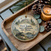 A decorative ornament featuring a historical scene with a cannon, boats, and a castle, placed on a wooden tray alongside pine cones and a lit candle.