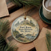 A round ornament with a picture of Fort Knox, Maine, featuring a cannon, a sailboat, and a building, is displayed on a wooden surface alongside a cup of coffee and some pine branches.