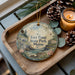 A decorative ornament with a scenic image of Fort Point State Park in Maine, featuring a castle, trees, and a river, is displayed on a wooden tray alongside pine cones and a lit candle.