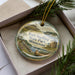 A decorative ornament in a box, featuring a scenic image of Fort Snelling, Minnesota, with a pine tree branch and greenery surrounding it.