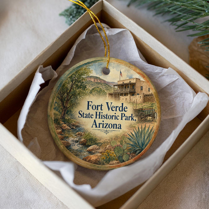 A round ornament with a painting of Fort Verde State Historic Park in Arizona, displayed in a box with a white tissue paper lining.