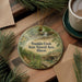 A round ornament with a scenic image of Franklin Creek, the State Natural Area in Illinois, is displayed on a wooden surface alongside a cup of coffee and some pine branches.