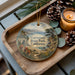 A decorative ornament featuring a scenic landscape of the coastal region of the state park in California, with a bridge and pine cones on a wooden tray.
