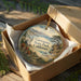 A decorative ornament in a wooden box, featuring a scenic landscape with mountains, a bridge, and a body of water.