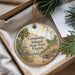 A round ornament with a lighthouse and palm trees, hanging from a string, placed inside a box with pine branches.