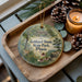 A decorative ornament with a forest scene and text, placed on a wooden tray next to pine cones and a lit candle.
