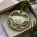 A decorative ornament in a box, featuring a scenic landscape with a waterfall and flowers.