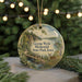 A round ornament with a scenic landscape featuring a bridge, deer, and trees, hanging in front of a Christmas tree with lights.