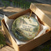 An ornament in a brown box, featuring a scenic landscape with a bridge, trees, and a deer.
